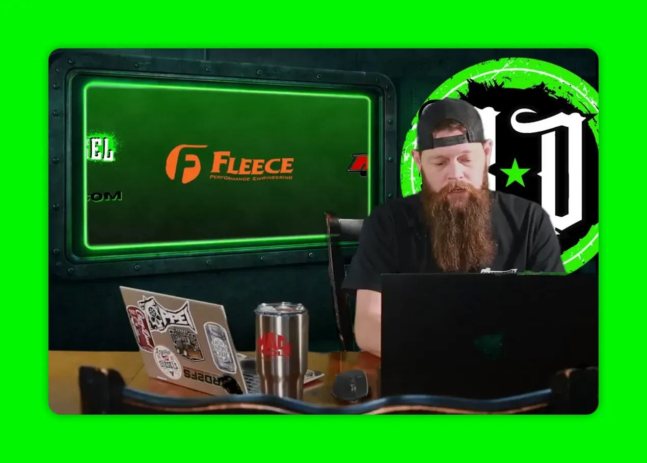 Presenter at a desk with a green display screen behind showing sponsor logos, clear mid-shot.